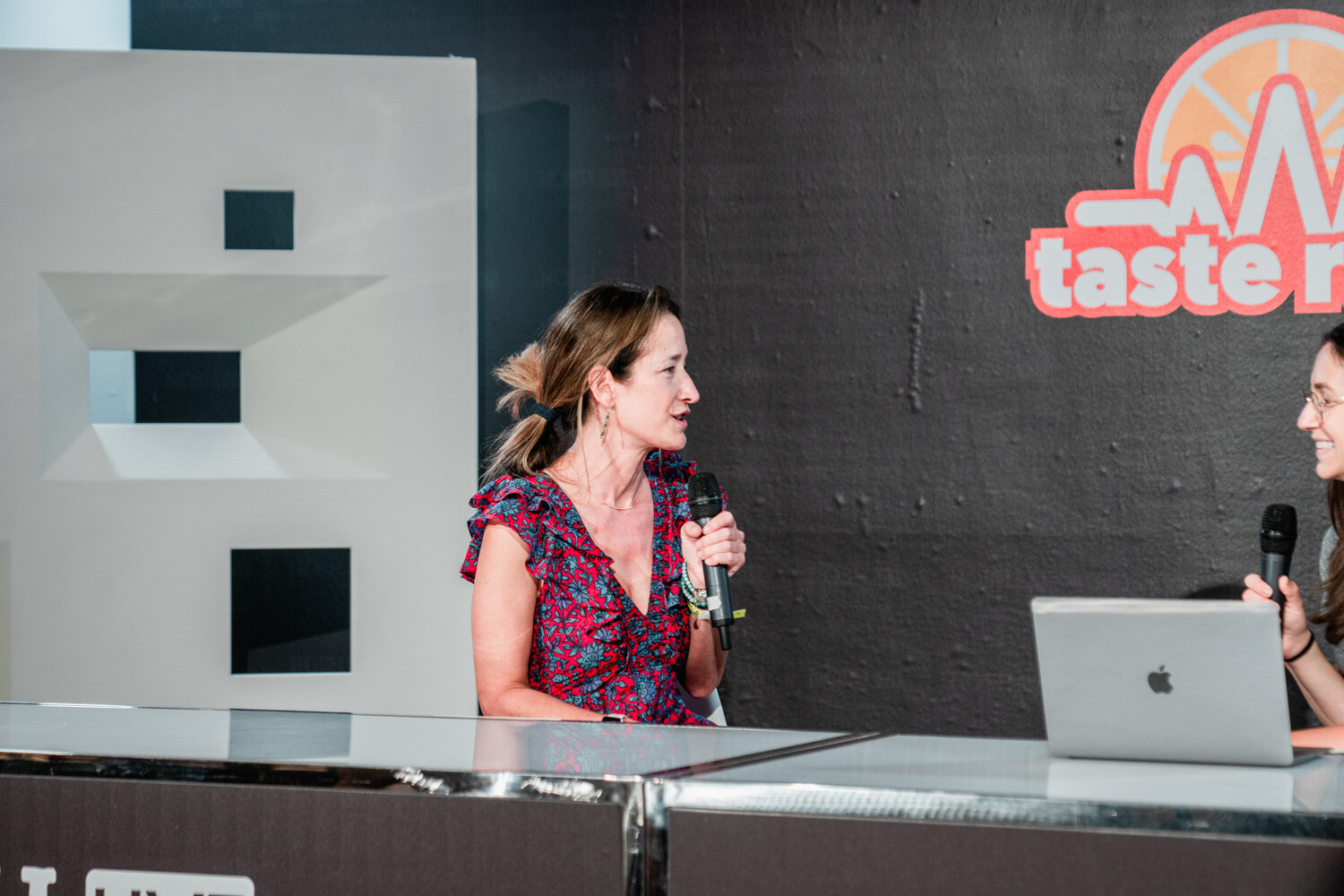 Woman in a red floral top holding a mic beside a laptop at the studio desk.