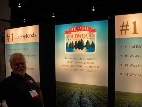A bearded man in a dark shirt and badge stands beside Wildwood Natural Foods display panels reading #1 in Soyfoods and a mission statement over a field image.