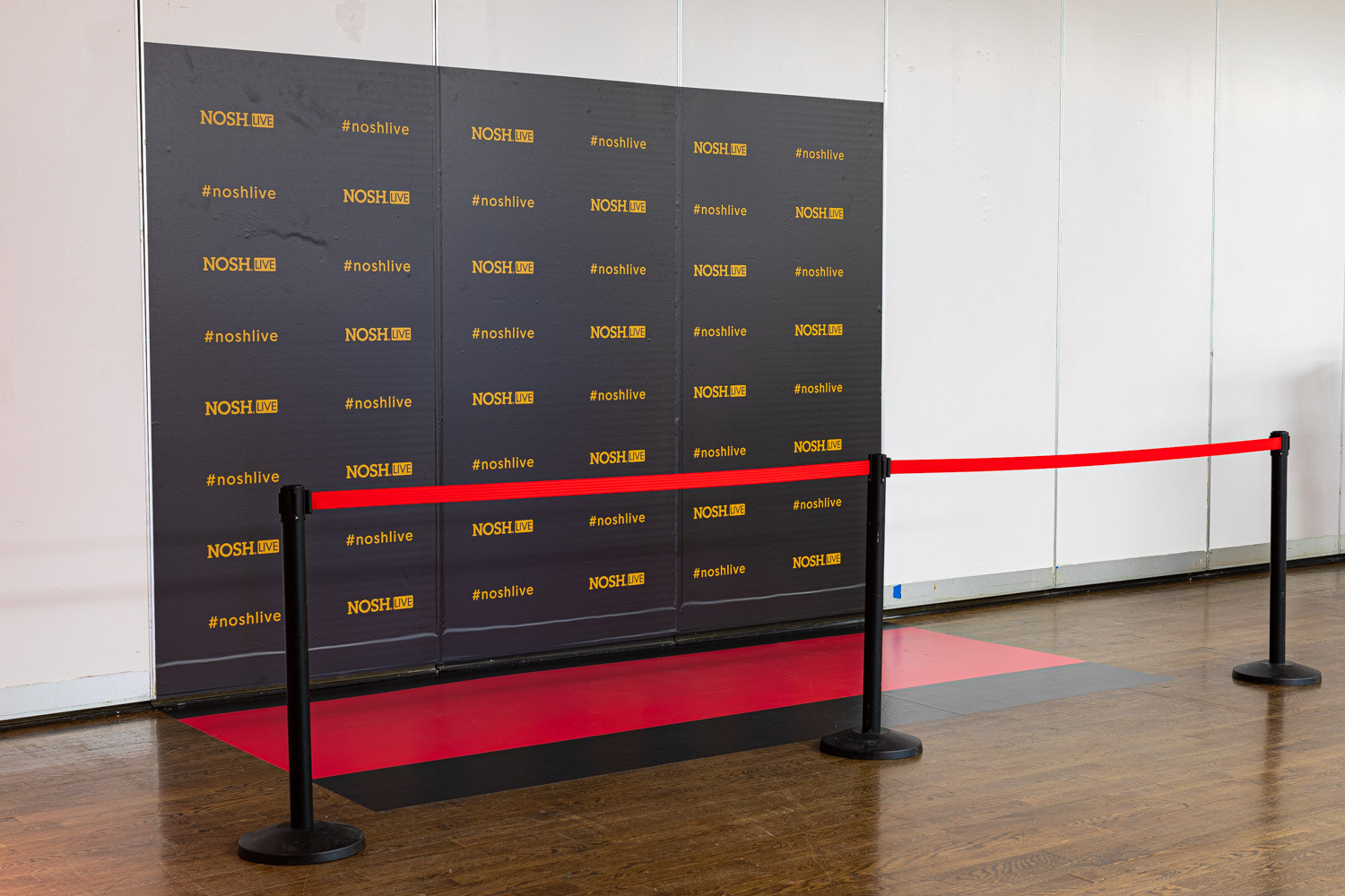 Black backdrop printed with Nosh Live and #noshlive logos, a red carpet and two stanchions with a red rope in front.