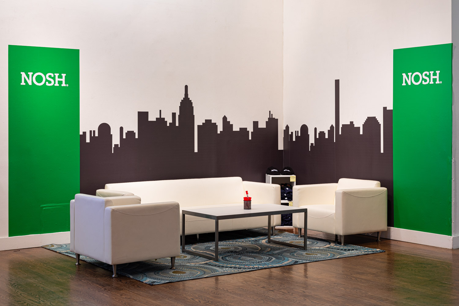White sofas and a coffee table on a patterned rug in a corner with green Nosh panels and a skyline silhouette.