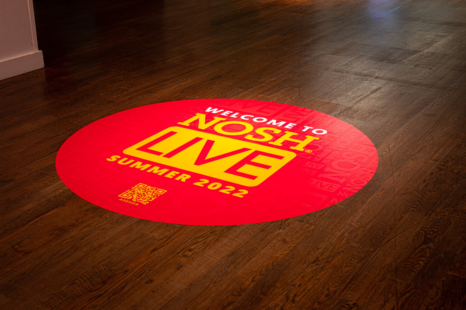 Round red decal on a dark wood floor reading Welcome to Nosh Live Summer 2022 with a QR code.