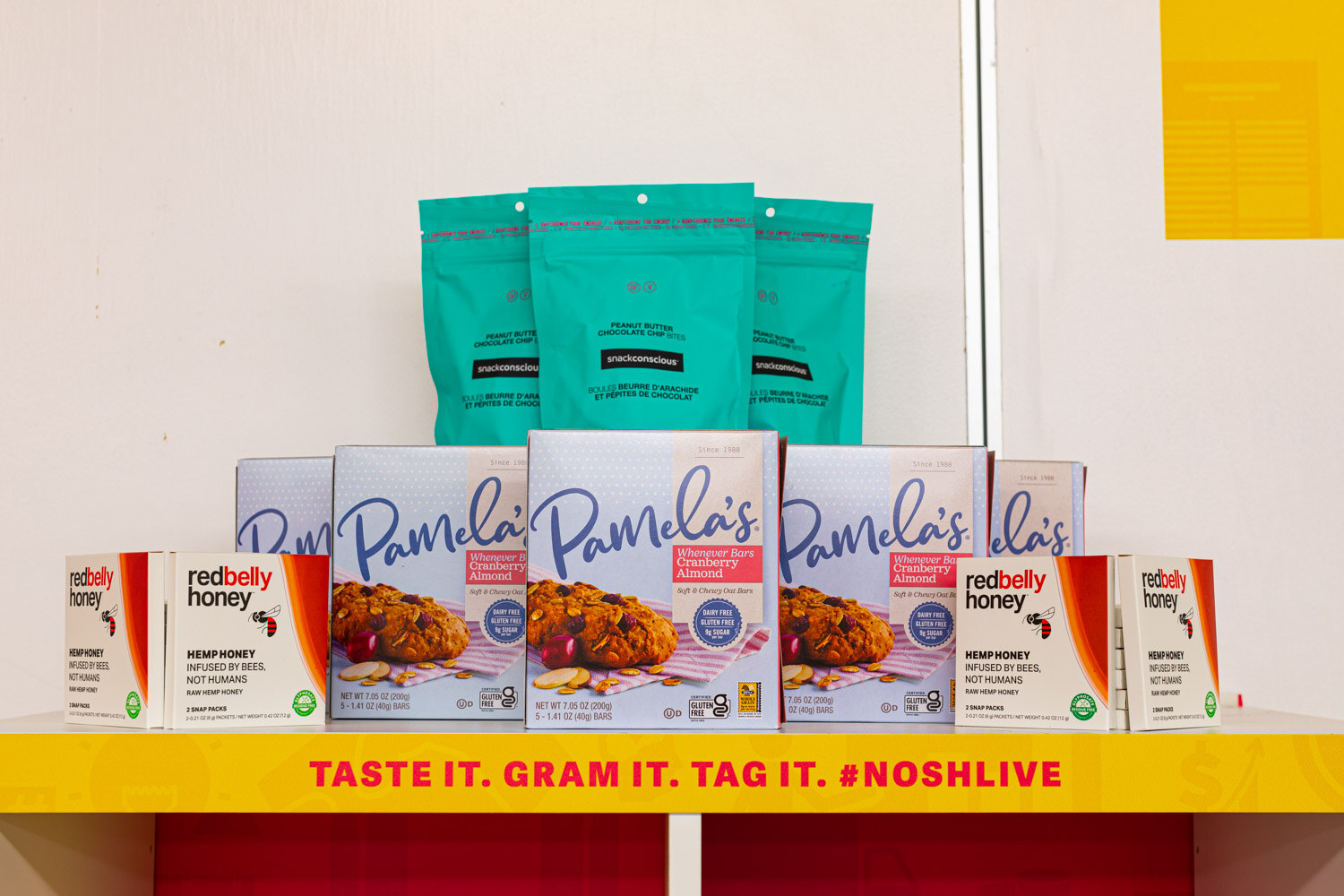 Boxes of Pamela's cookies and Red Belly Honey in front of teal pouches on a yellow shelf reading Taste it. Gram it. Tag it. #noshlive.