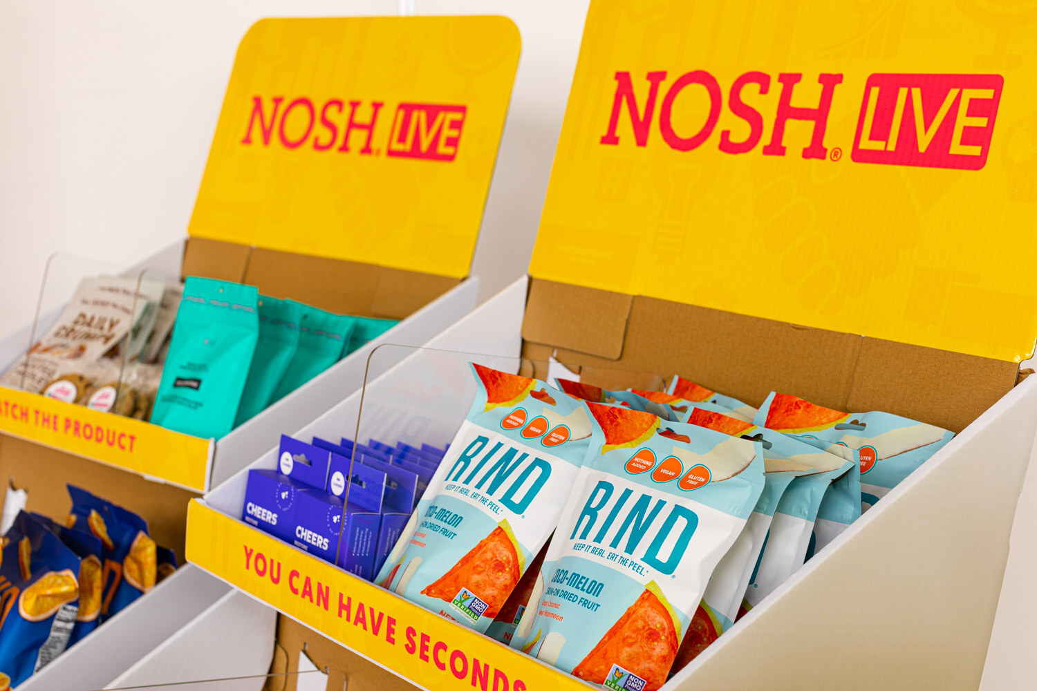 Two yellow Nosh Live cardboard display bins holding Rind sea melon bags, Cheers packets and other pouches.