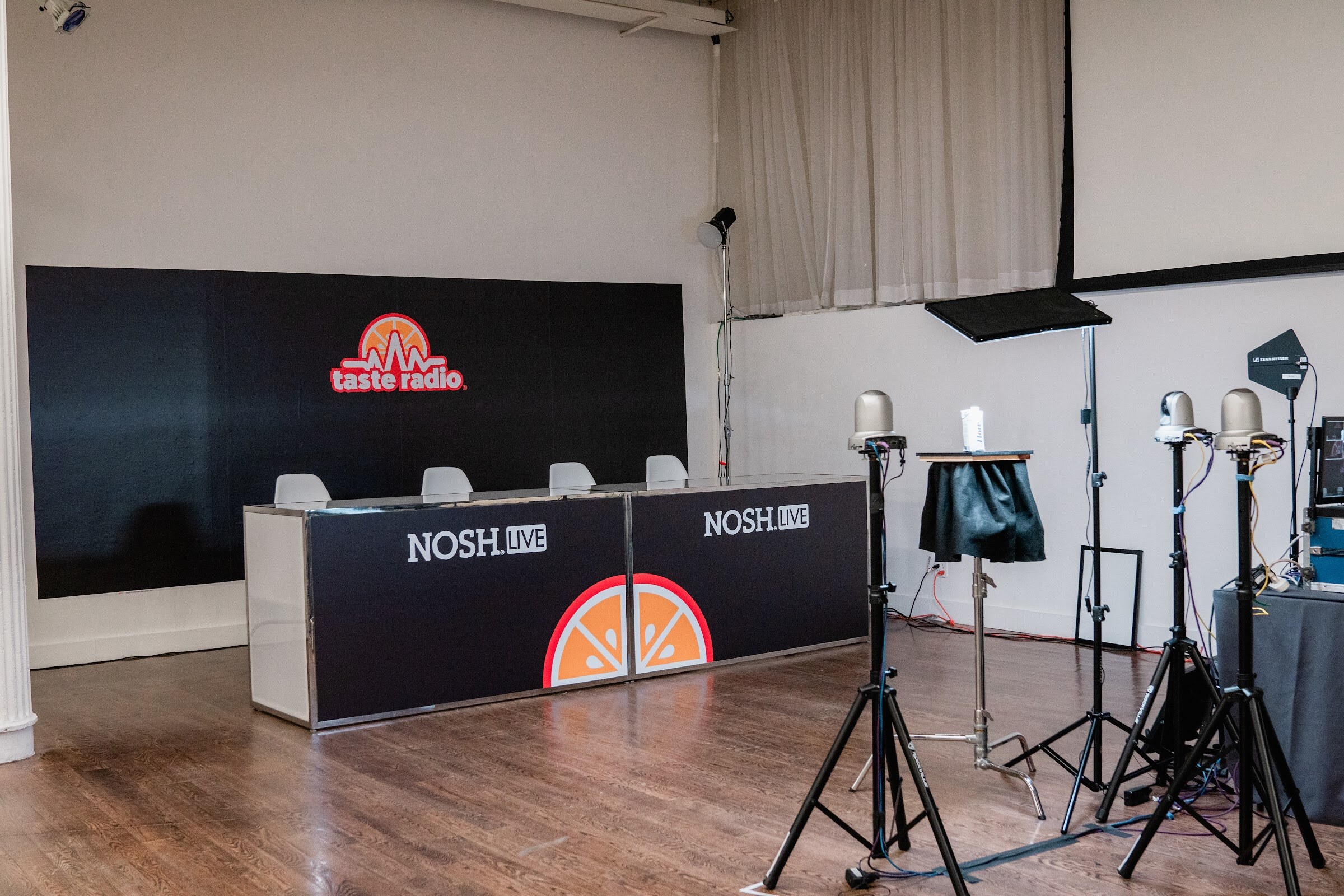 Black Nosh Live-branded desk in front of a Taste Radio wall, with lighting stands and a camera stand on the wood floor.
