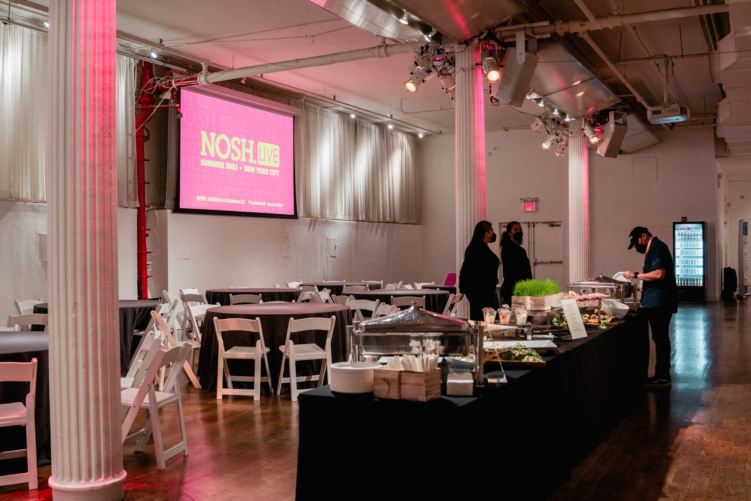 Long black buffet table with salads and staff at the right, round tables and a pink Nosh Live screen on the wall.
