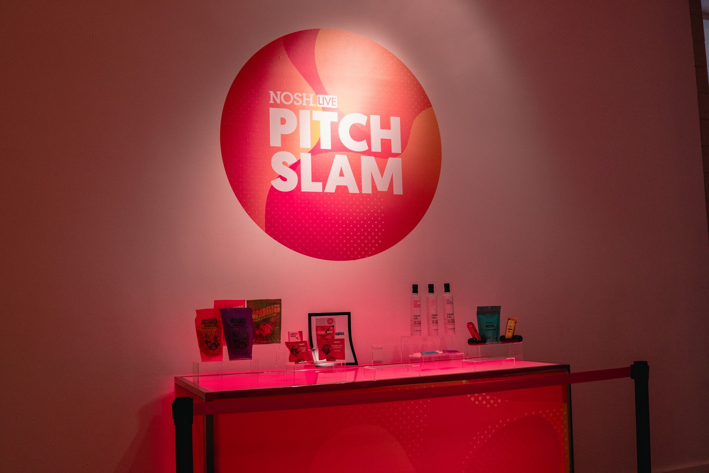 Round Pitch Slam logo on a red-lit wall above a table with products and bottles behind a red rope.