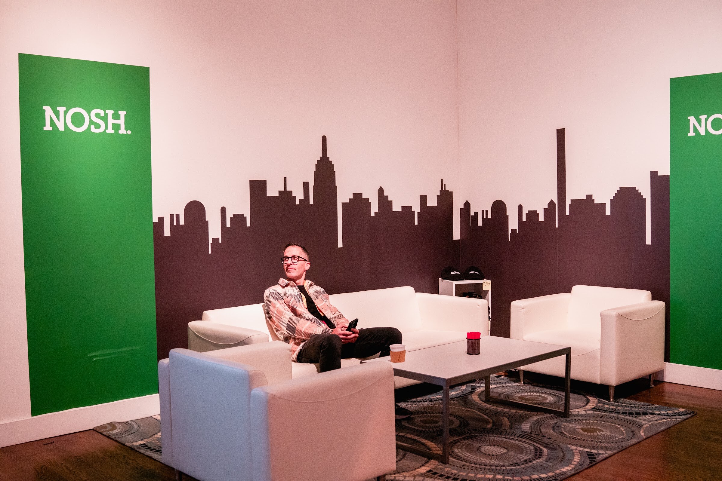Man in a plaid jacket seated on a white sofa in front of a New York skyline graphic and green Nosh panels.