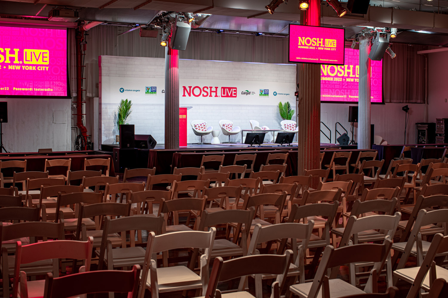 Wide view of empty folding chairs facing a stage with a Nosh Live backdrop and pink screens hung from the ceiling.