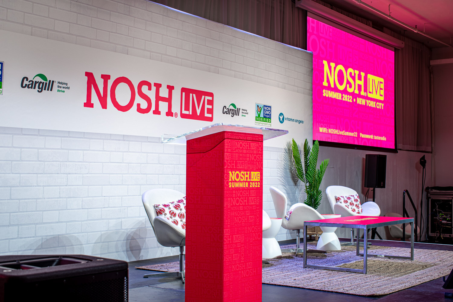 Red podium reading Nosh Live Summer 2022 beside white chairs with patterned pillows, a Nosh Live wall and a pink screen.