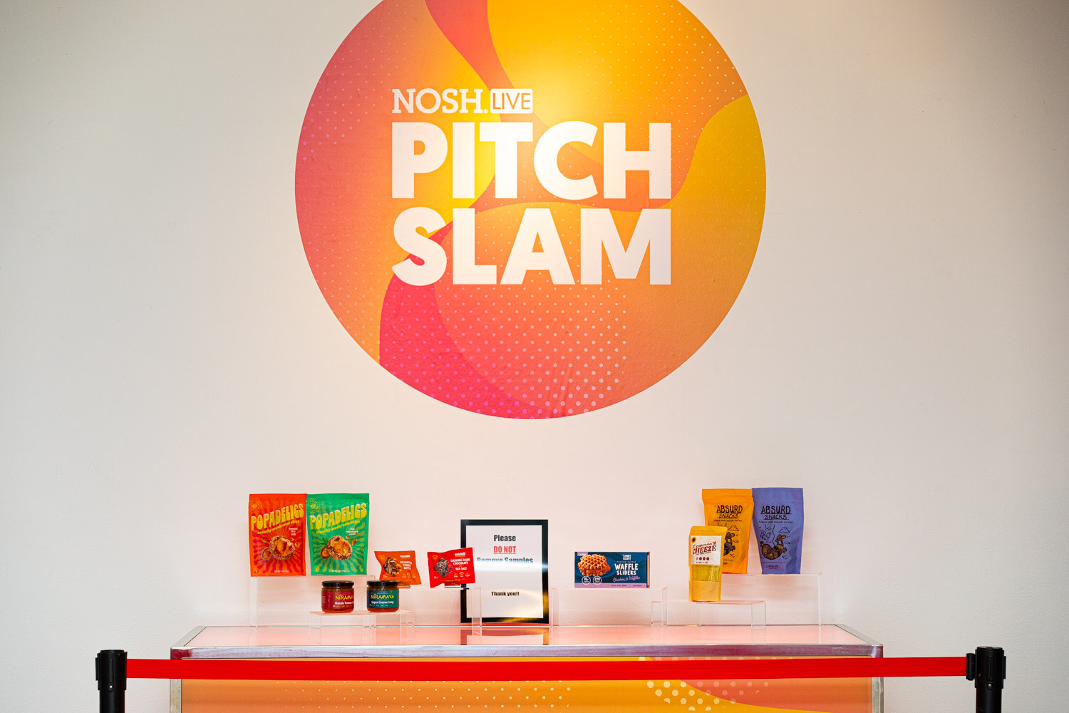 Orange Pitch Slam logo on a white wall above a lit table with pouches, jars and packages behind a red rope.