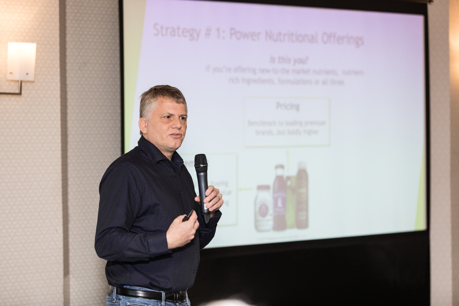 A man in a dark shirt holds a microphone beside a projection screen titled Strategy 1, Power Nutritional Offerings, showing pricing text and bottles of juice.