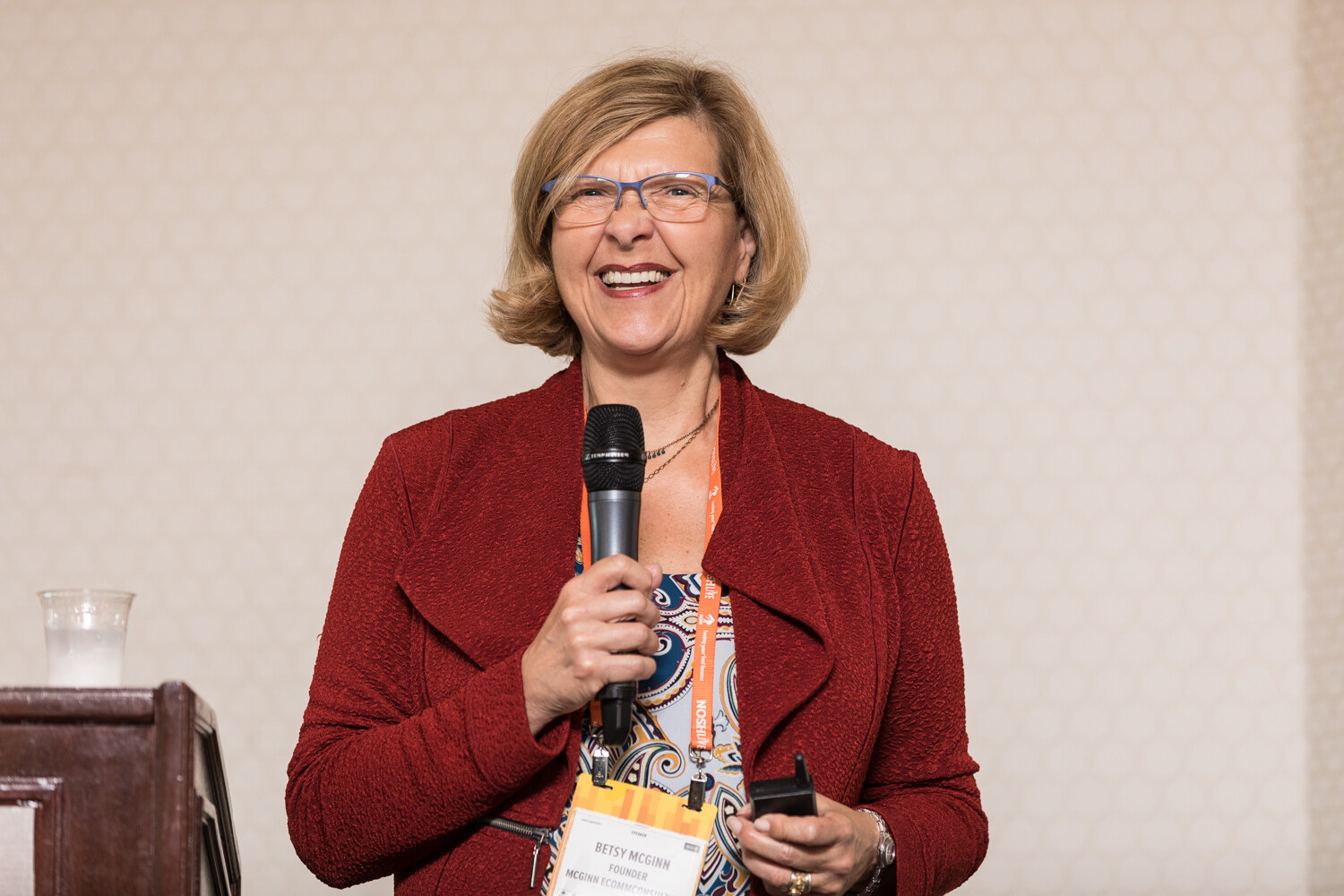 A smiling woman in a red jacket holds a handheld microphone and a slide clicker in front of a beige wall.