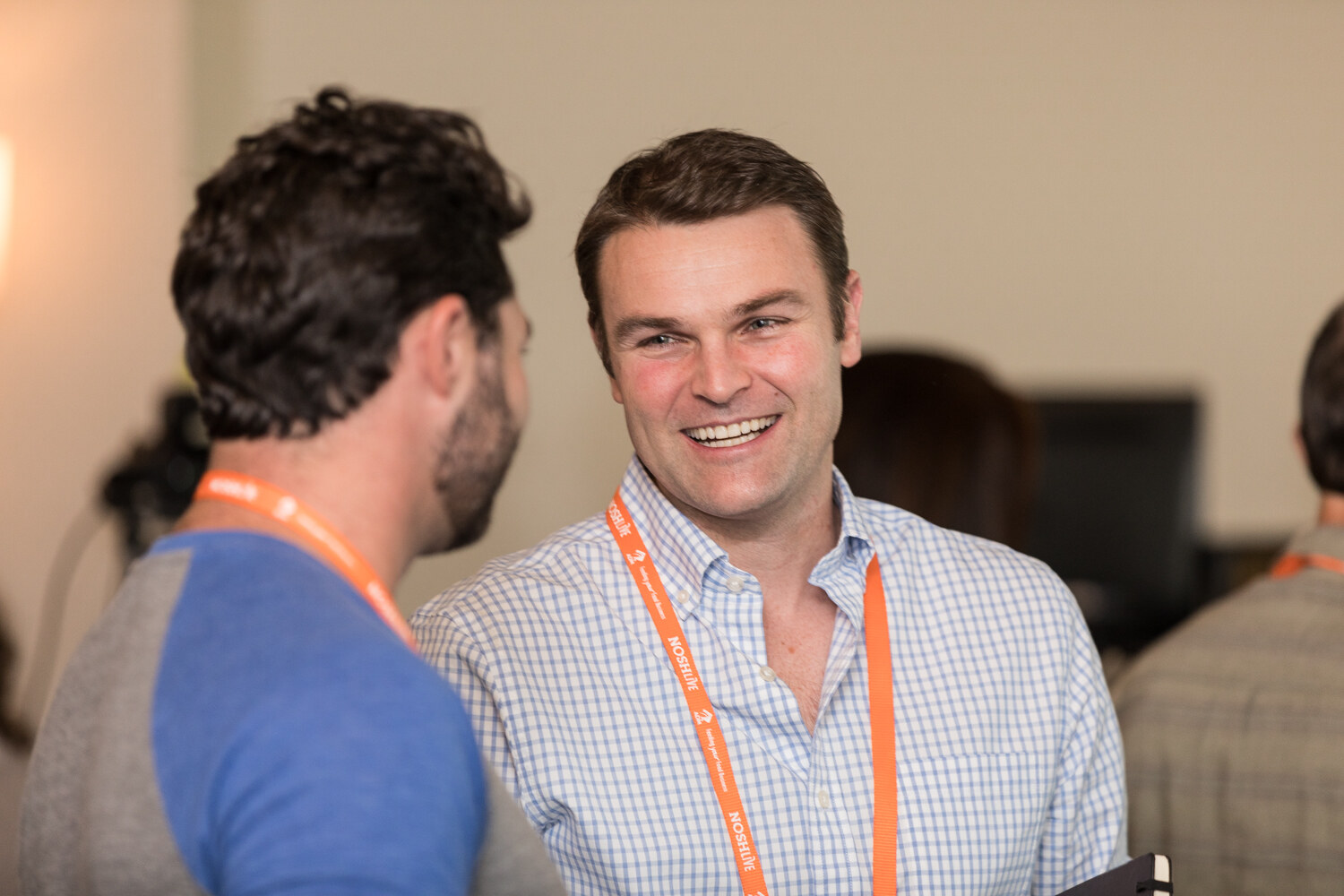 A smiling man in a light blue checked shirt with an orange NOSH Live lanyard talks with a man in a gray and blue shirt, seen from behind.