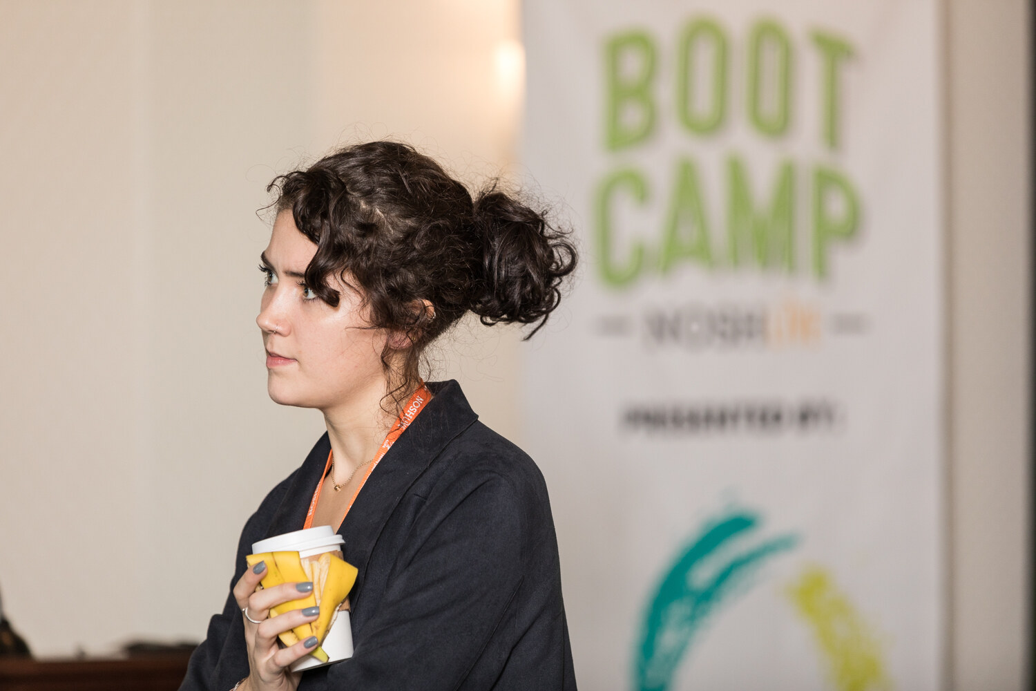 A woman with a curly updo and a dark jacket holds a coffee cup in front of a banner reading Boot Camp NOSH Live, presented by.