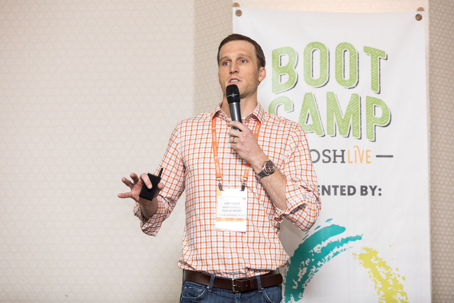 A man in an orange checked shirt holds a microphone and a slide clicker in front of a banner reading Boot Camp NOSH Live.