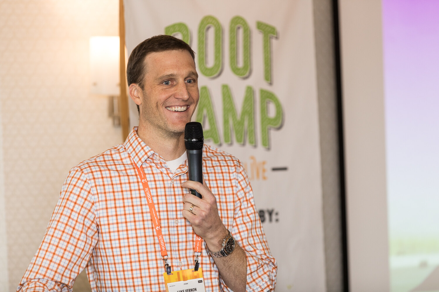 A smiling man in an orange checked shirt holds a handheld microphone in front of a Boot Camp banner.