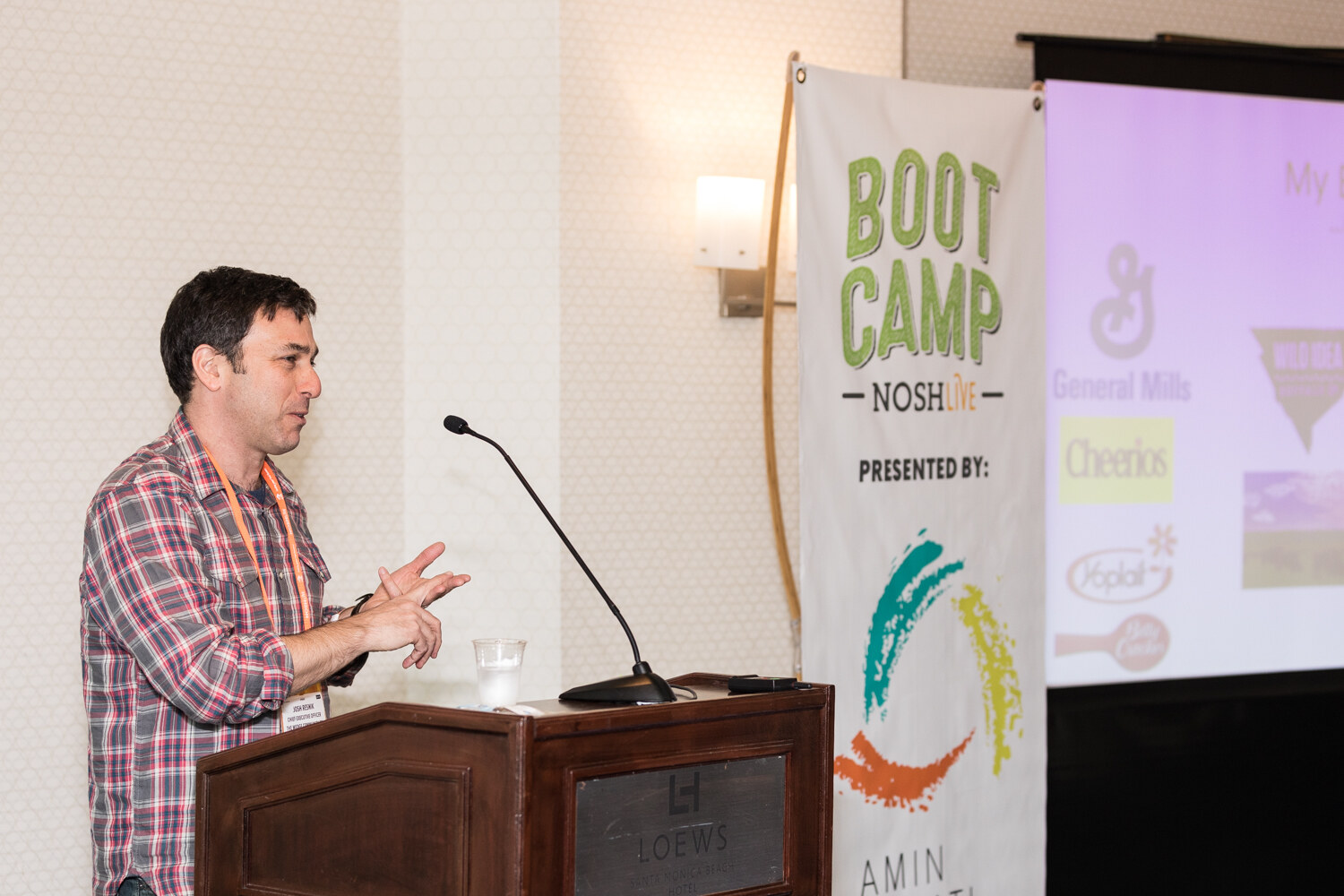 A man in a red plaid shirt speaks at a wooden lectern marked Loews, next to a Boot Camp NOSH Live banner and a projected slide with General Mills, Cheerios and Yoplait logos.