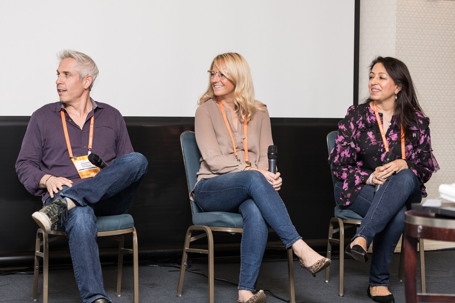 Three people sit in a row of chairs holding microphones in a small meeting room, with a blank projection screen behind them; two look to the left.