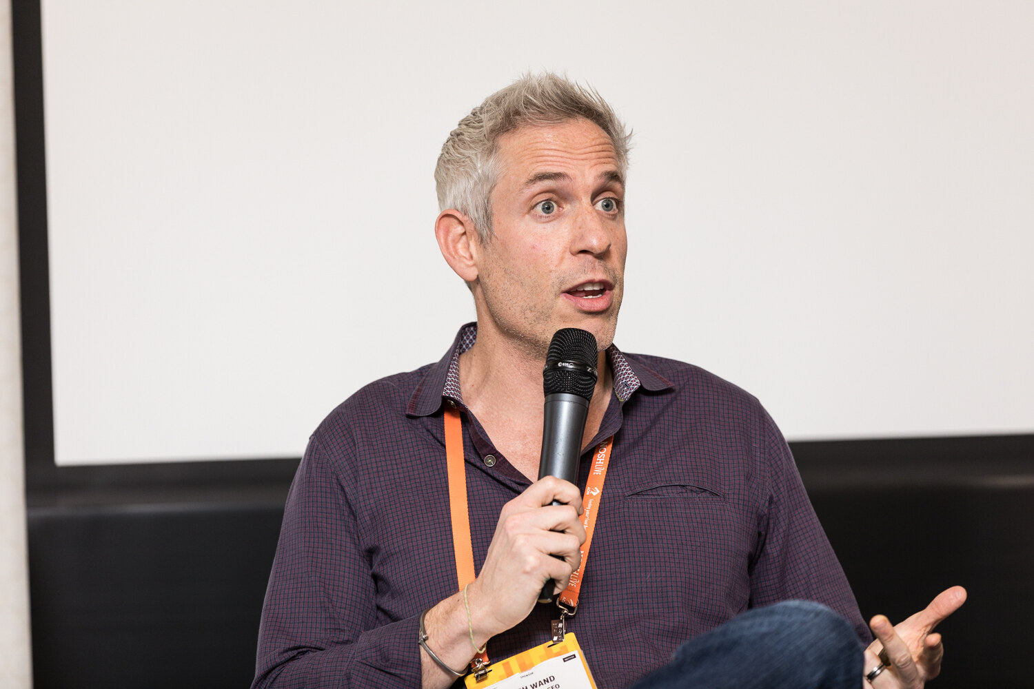 A gray-haired man in a dark checked shirt speaks into a handheld microphone and gestures with one hand in front of a projection screen.