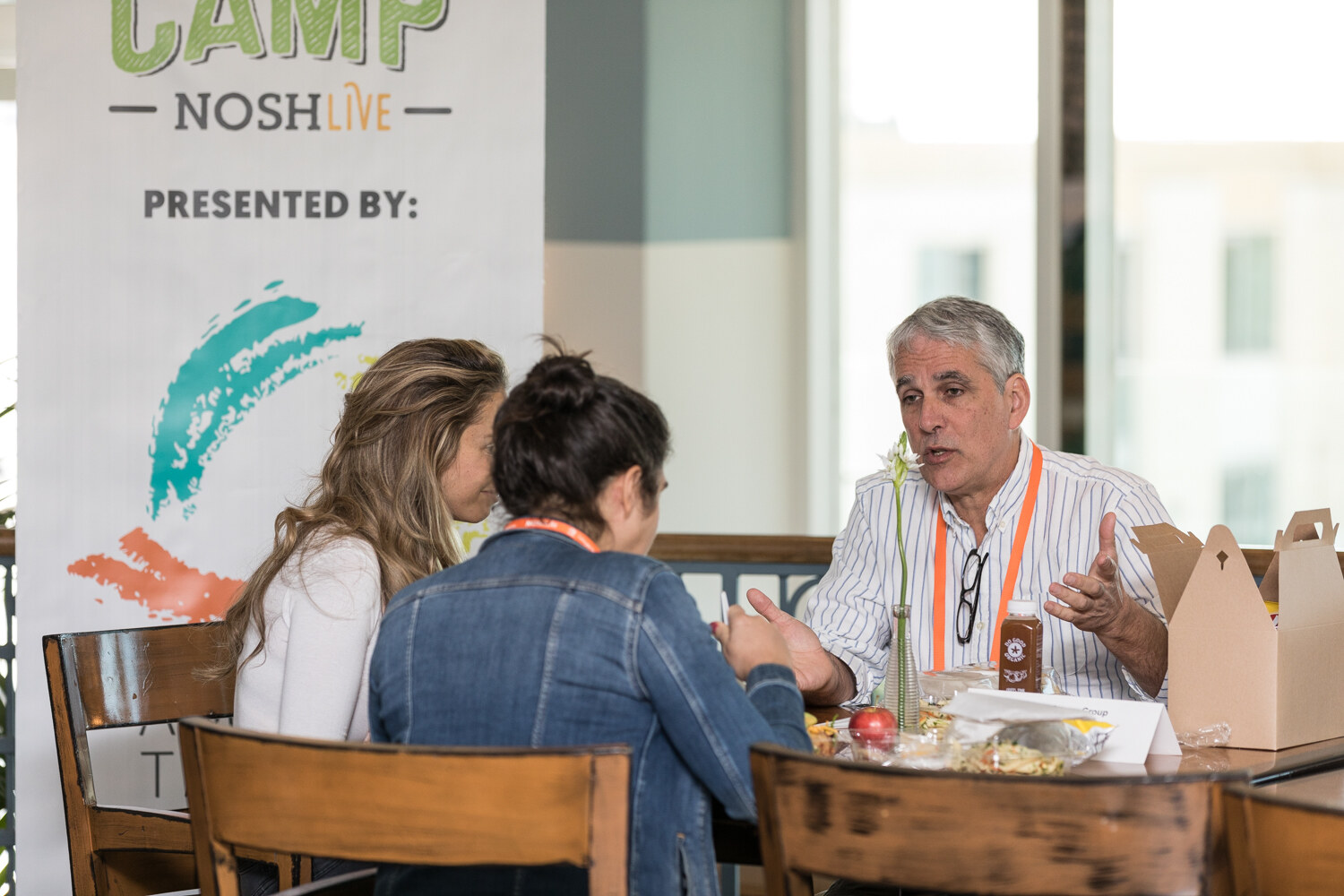 A gray-haired man in a striped shirt gestures while talking with two women at a wooden table near a banner reading Boot Camp NOSH Live, presented by.