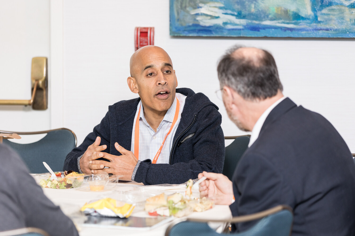 A bald man in a navy zip-up jacket and orange lanyard gestures while talking to a man in a dark blazer seen from behind, with salads on the table.