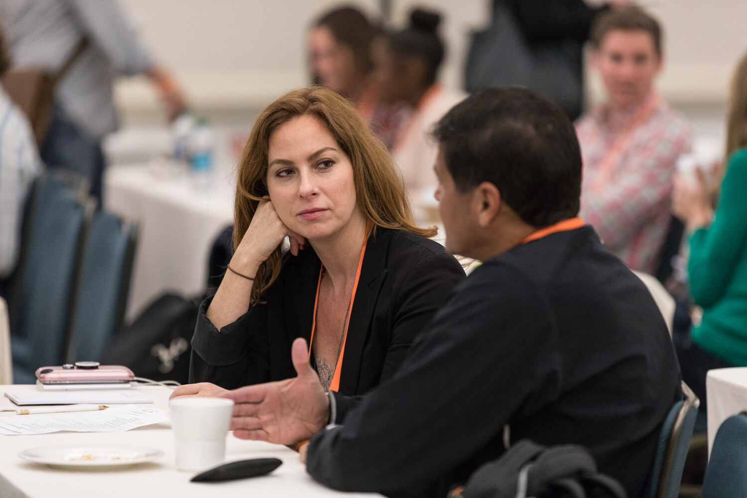 A woman in a black blazer rests her chin on her hand as she listens to a man in a dark jacket in the foreground, at a table with a coffee cup and notes.
