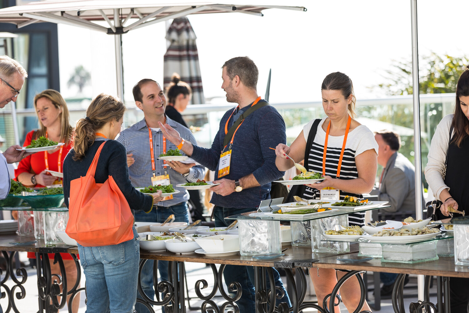 A group of attendees with orange lanyards hold plates and serving spoons along a long outdoor buffet table under a patio umbrella; a woman in the foreground carries an orange tote bag.