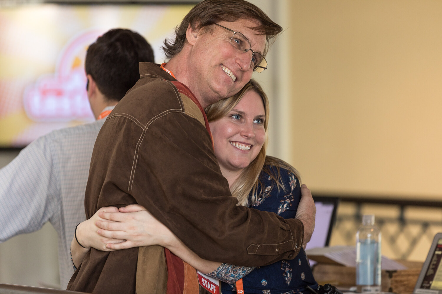 A smiling man in glasses and a brown jacket hugs a smiling woman in a floral top, with a staff badge visible at the bottom.