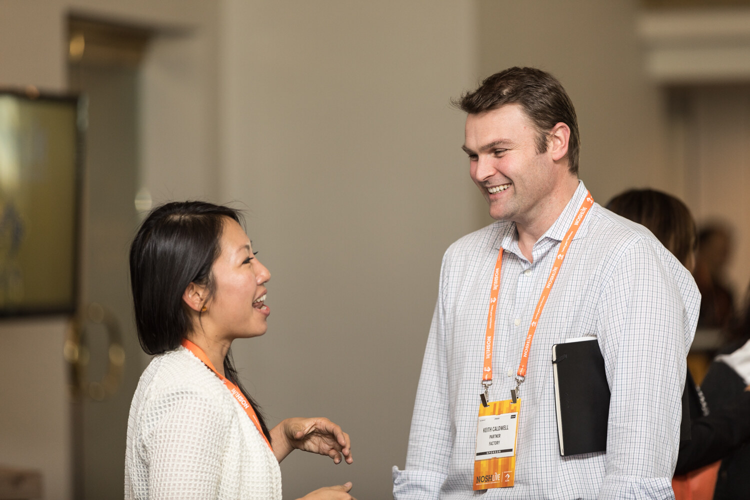 A woman in a white cardigan talks with a smiling man in a light checked shirt wearing an orange NOSH Live lanyard and holding a black notebook.