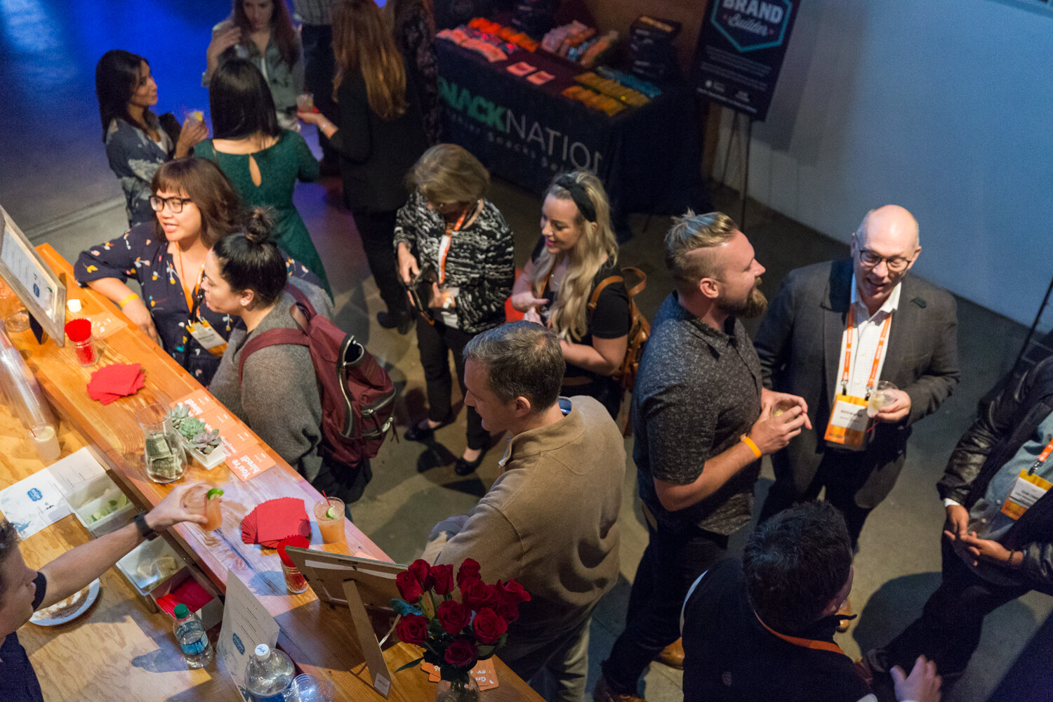 A high-angle view of guests with orange lanyards standing around a wooden bar with red napkins and roses, near a table with a Snack Nation banner and a Brand Builder sign.