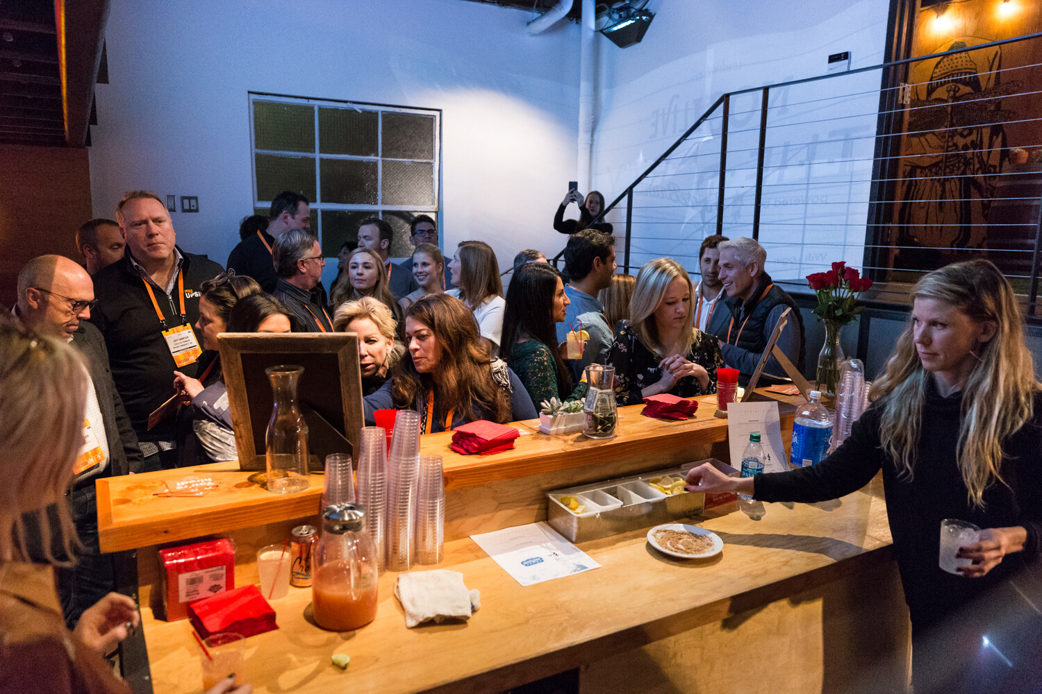 A busy wooden bar with plastic cups, a juice dispenser and a vase of red roses, with guests behind it and a woman in black reaching for a cup on the right.