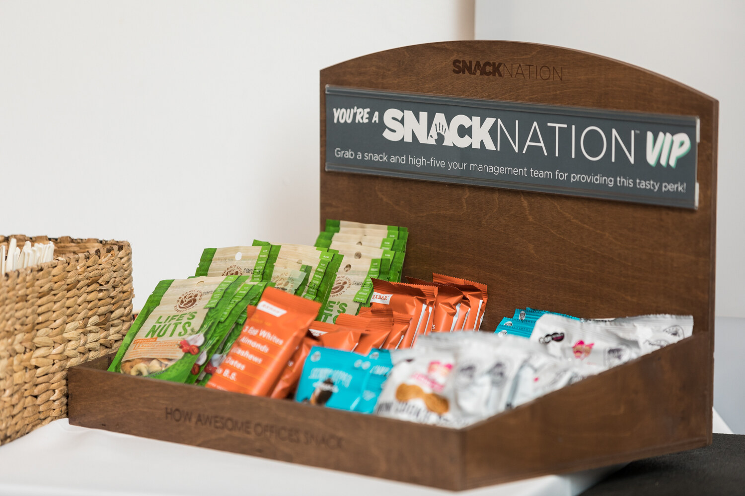 A wooden tray with a sign reading You're a Snack Nation VIP holds pouches of nuts, almond bars and other snacks beside a woven basket.