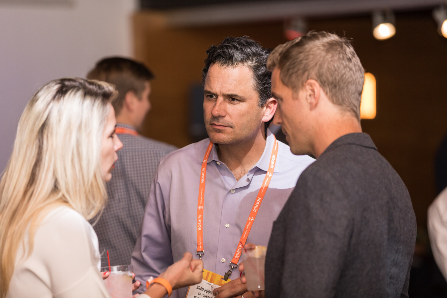 A man in a lavender shirt with an orange lanyard talks with a blonde woman and a man in a gray jacket, with warm lights behind them.
