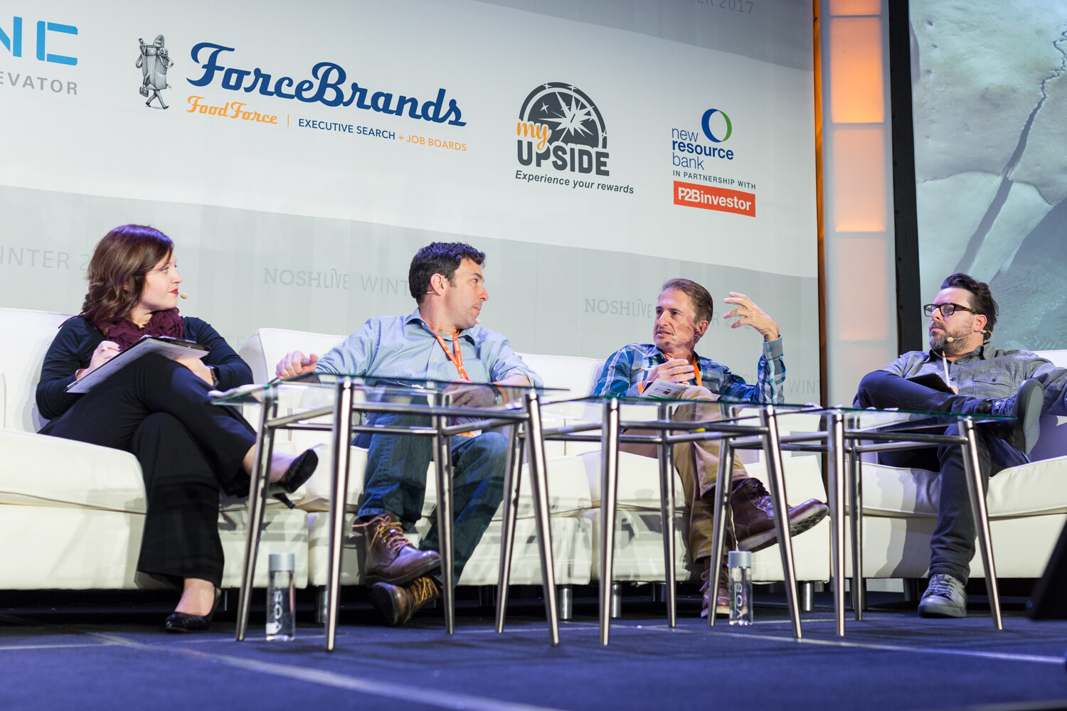 Four people sit on white sofas on a stage beneath ForceBrands, My Upside and New Resource Bank logos, talking among themselves.