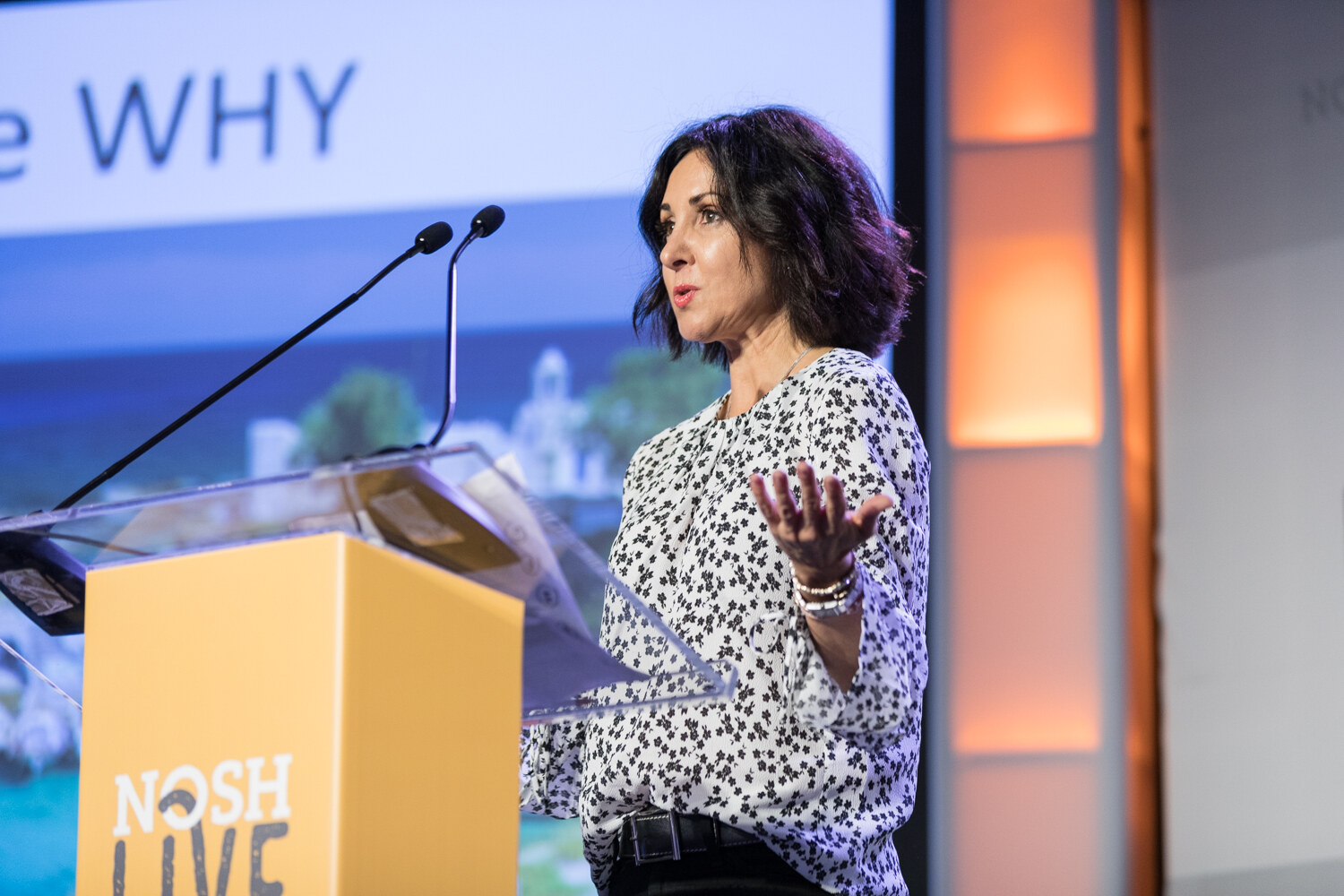 A dark-haired woman in a black and white patterned blouse gestures with one hand at a yellow NOSH Live podium in front of a slide reading the WHY.