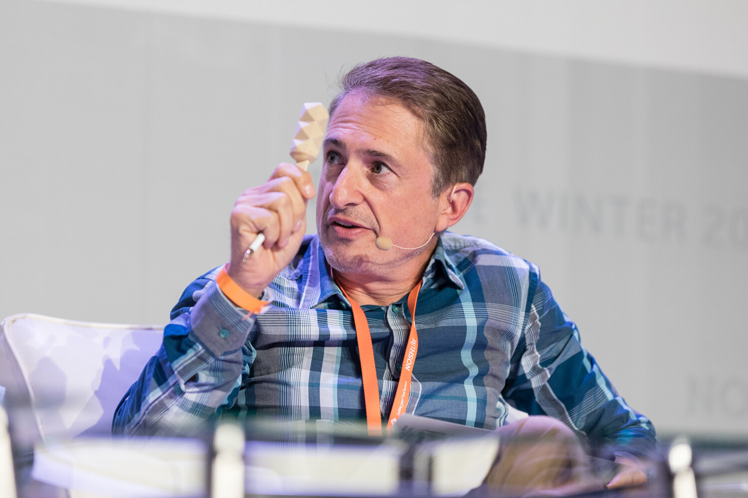 A man in a blue plaid shirt with a headset microphone holds up a small cream-colored geometric pop on a stick.