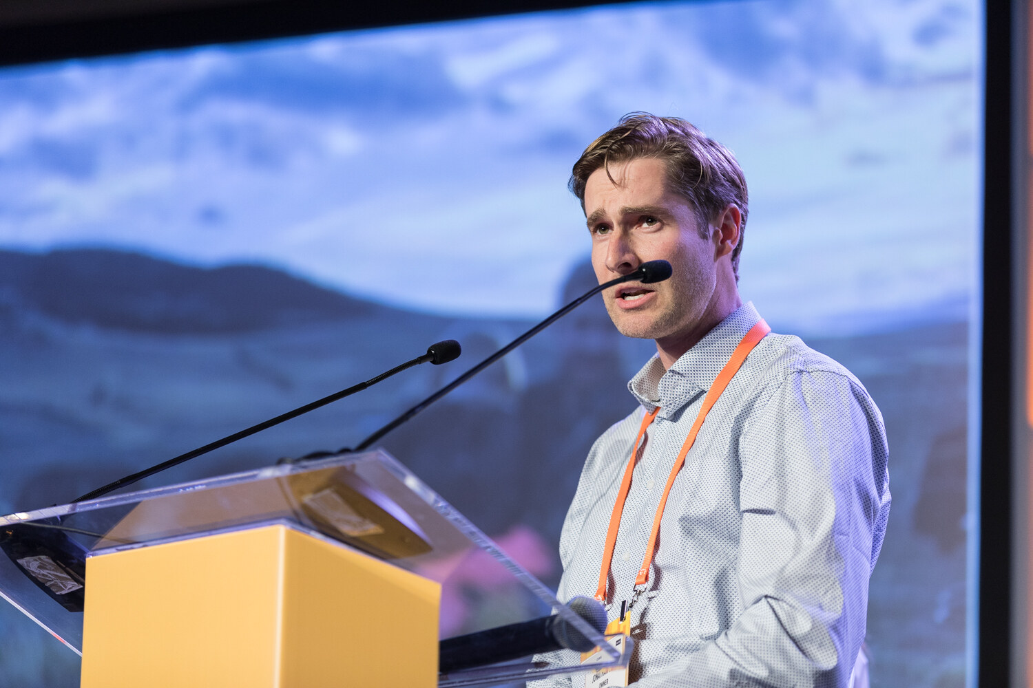 A young man in a light checked shirt with an orange lanyard speaks at a yellow podium with two microphones in front of a slide of hills and sky.