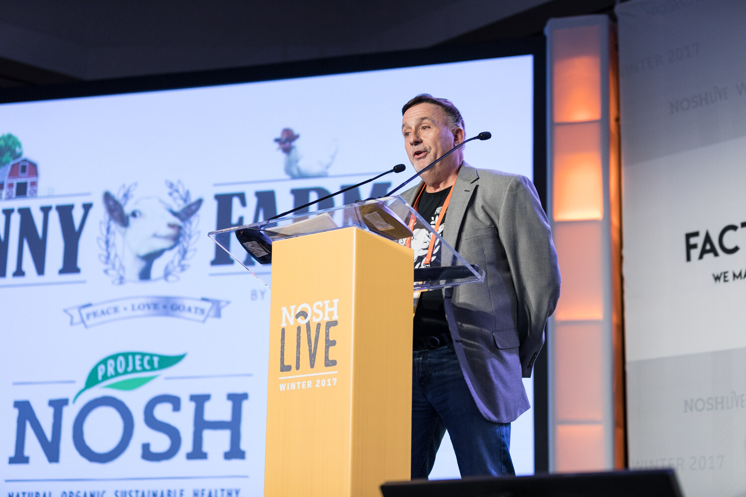A man in a gray blazer stands at a yellow podium in front of a large slide with a goat illustration and the Project NOSH logo.