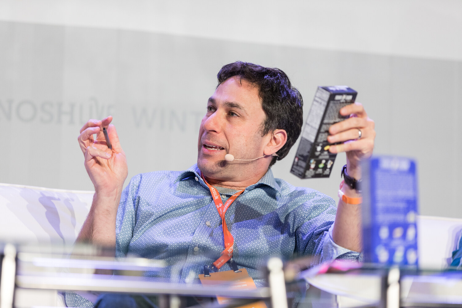 A dark-haired man in a blue shirt with a headset microphone holds a black product box in one hand and a pen in the other.