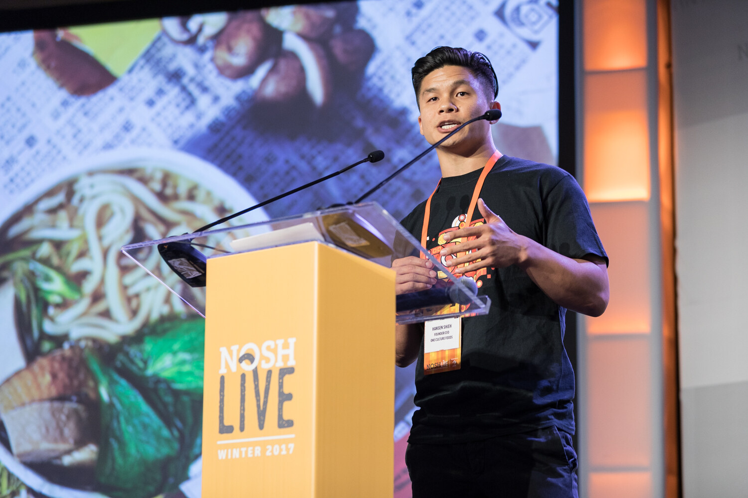 A young man in a black T-shirt with an orange lanyard speaks at the NOSH Live Winter 2017 podium in front of a slide showing a bowl of noodles with mushrooms and greens.