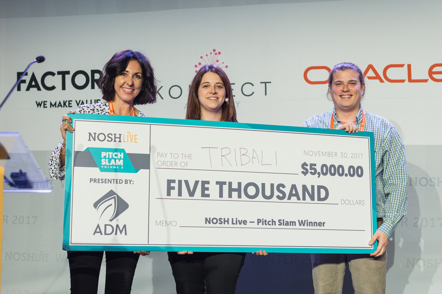 Three people on stage hold an oversized check reading NOSH Live Pitch Slam, presented by ADM, pay to the order of Tribali, five thousand dollars, dated November 30, 2017.