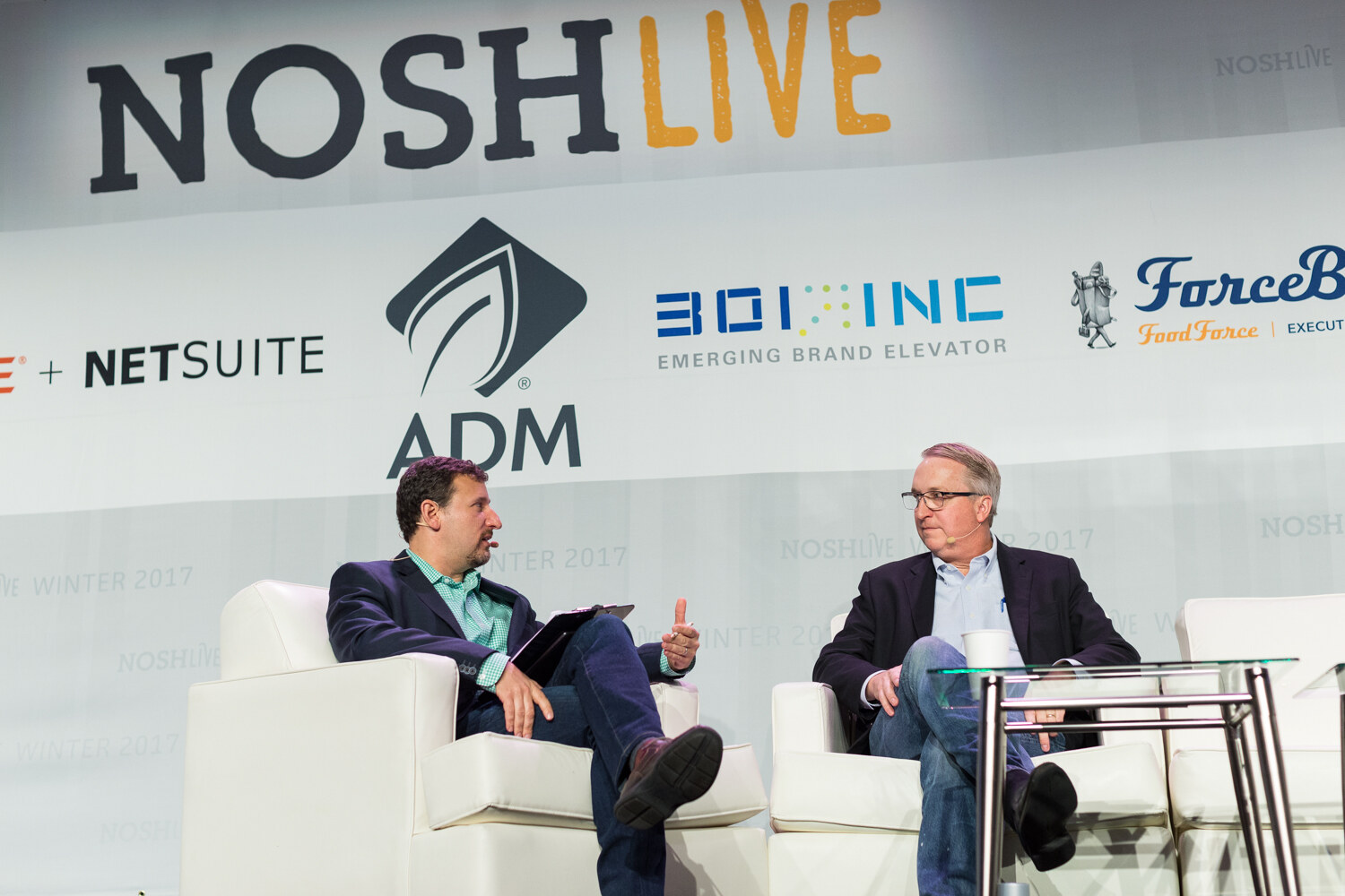 Two men in blazers talk in white armchairs on a stage under a NOSH Live banner with ADM, 301 Inc and ForceBrands logos.