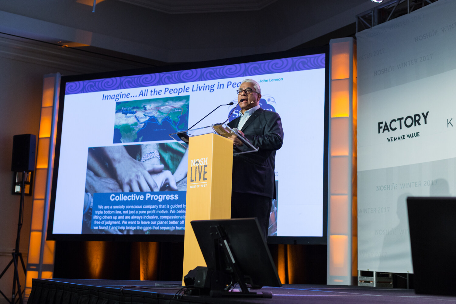 A man at a NOSH Live podium stands in front of a large screen showing a slide titled Collective Progress, next to a Factory banner.