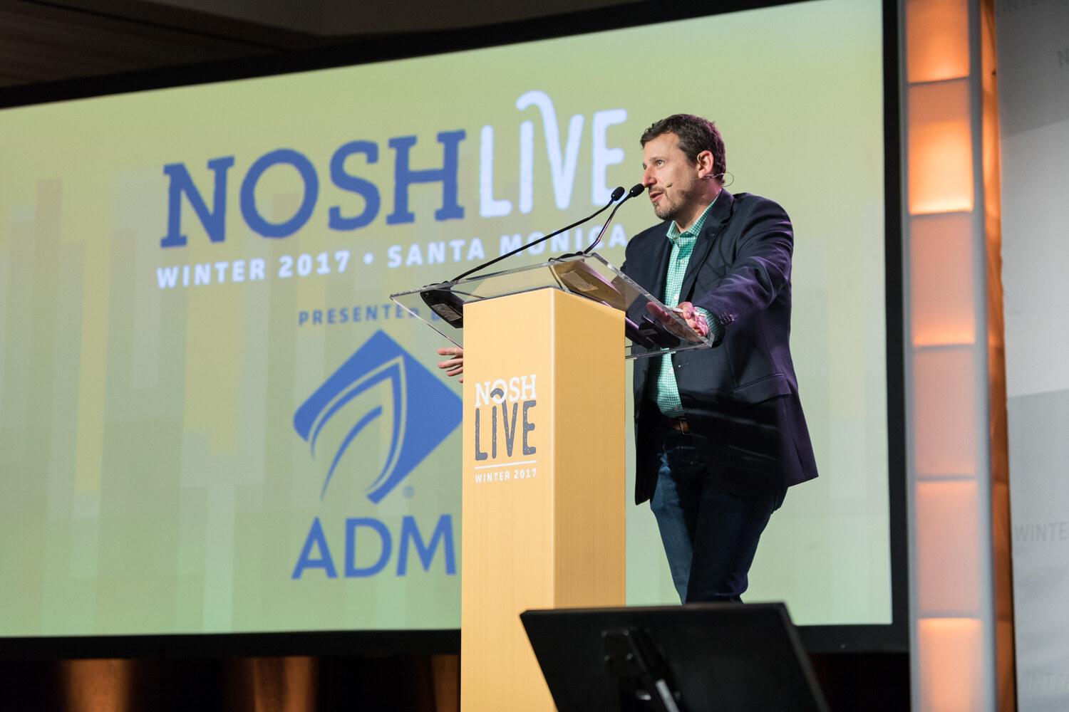 A man in a navy blazer and green checked shirt speaks at a yellow NOSH Live podium in front of a large screen reading NOSH Live Winter 2017, Santa Monica, presented by ADM.