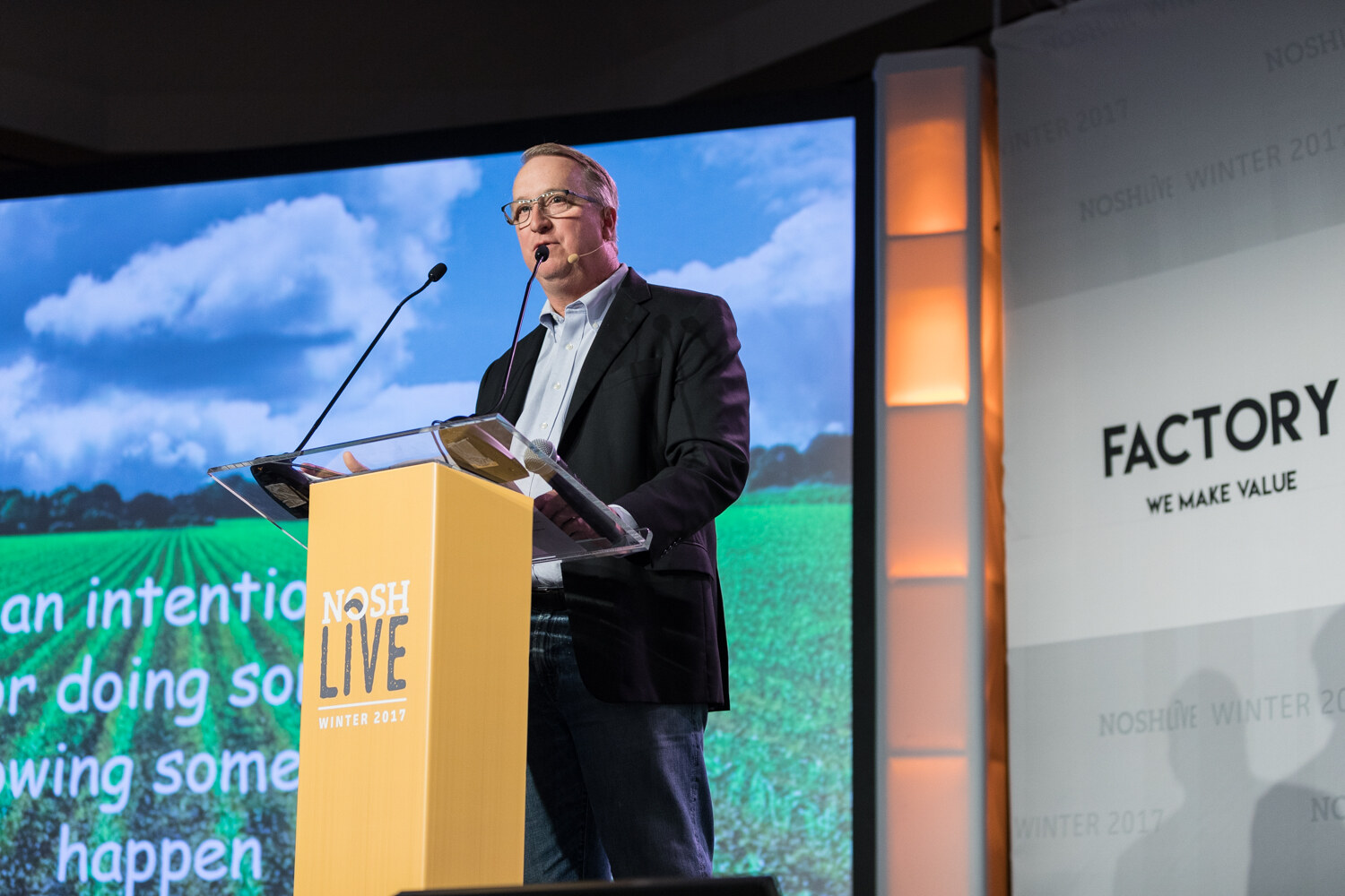 A man with glasses speaks at a yellow NOSH Live podium with two microphones, in front of a slide showing a green crop field.