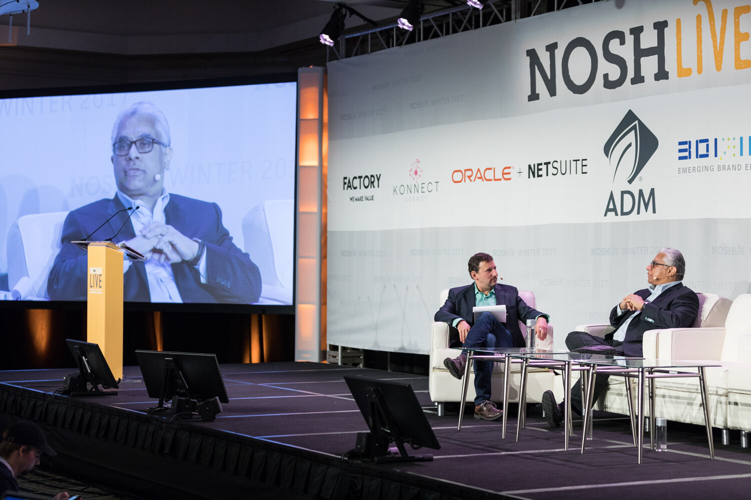 Two men sit in white chairs on a wide stage under a NOSH Live backdrop with sponsor logos, while a large screen at left shows a close-up of one of them.