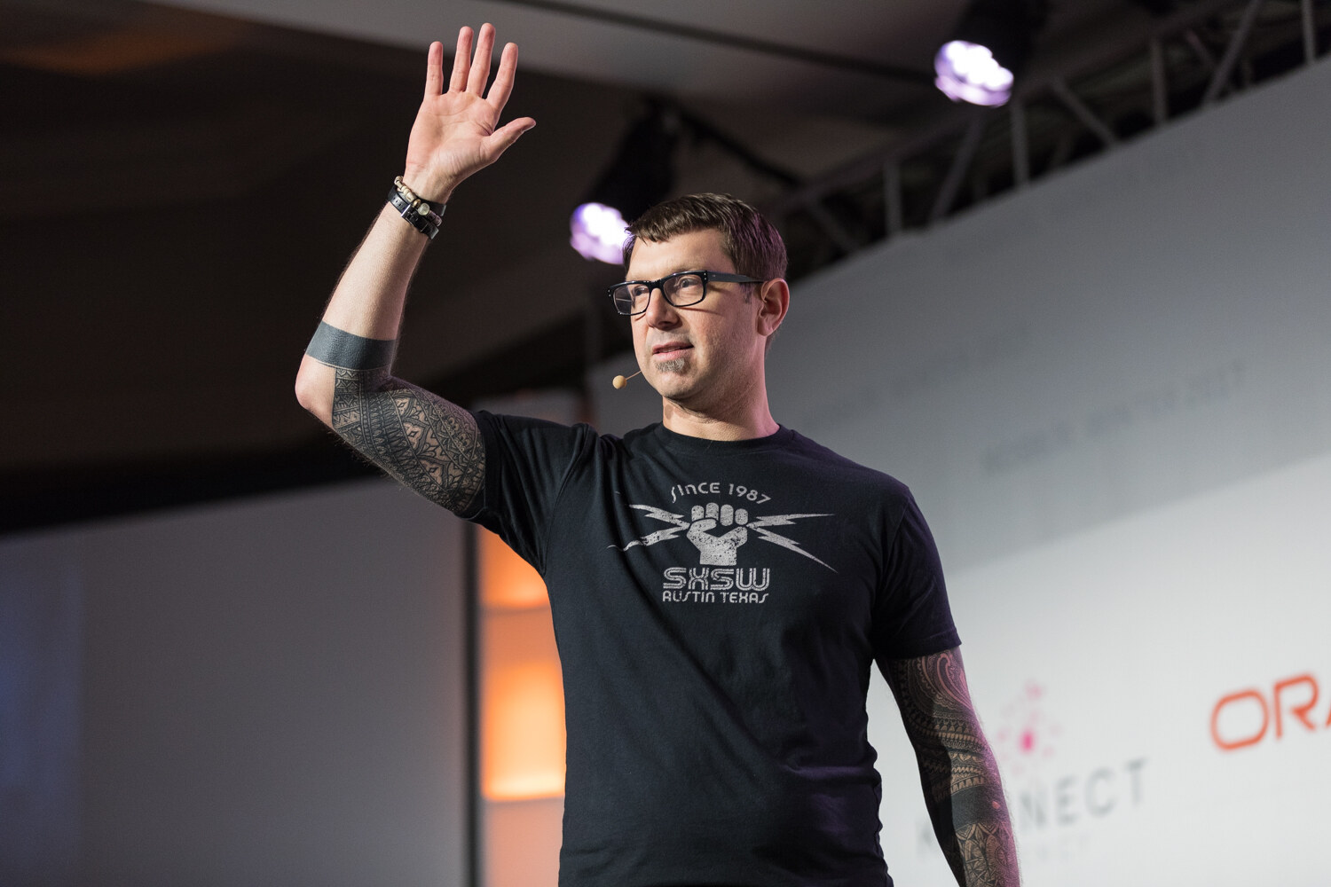 A man in a black SXSW T-shirt with tattooed forearms raises one hand on stage under spotlights.