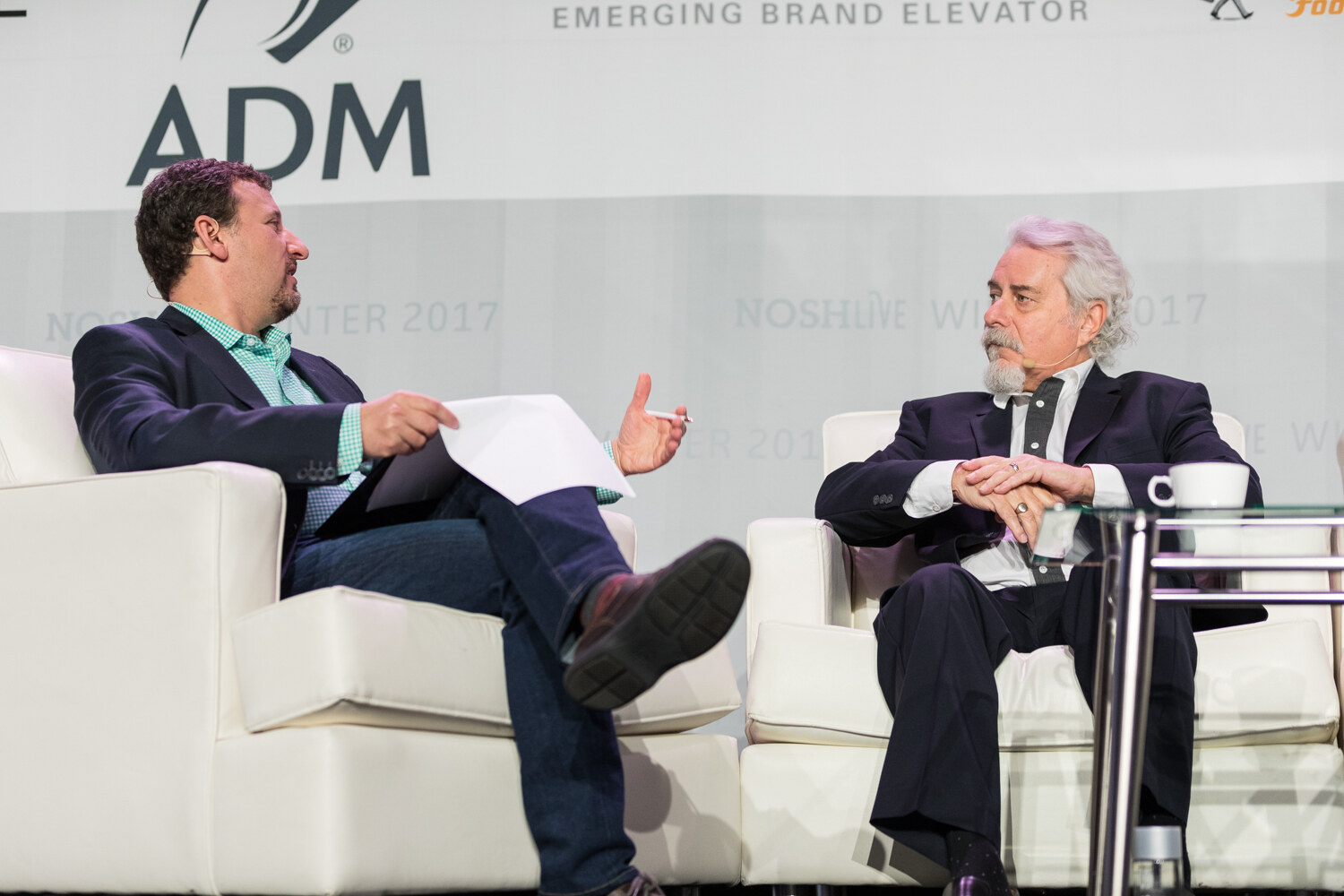 Two men face each other in white armchairs on the NOSH Live stage, with a coffee cup on the glass table and an ADM banner above.