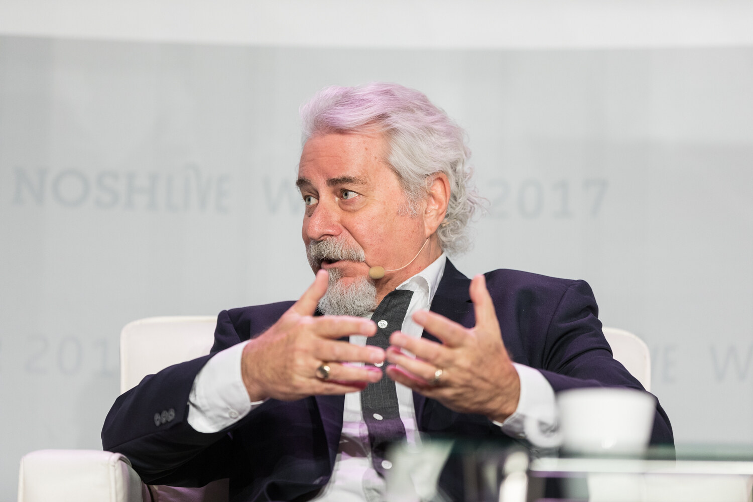 A gray-haired man with a goatee and a headset microphone gestures with both hands while seated.