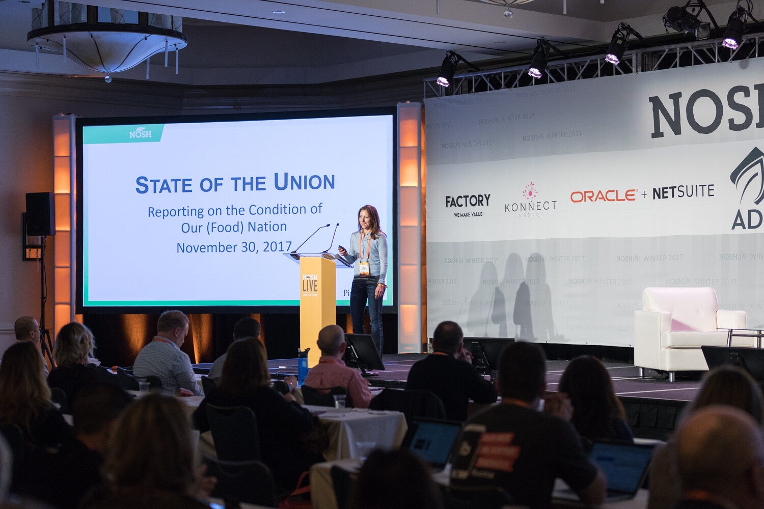 A woman at a yellow NOSH Live podium beside a projected slide titled State of the Union, with seated attendees with laptops in the foreground.